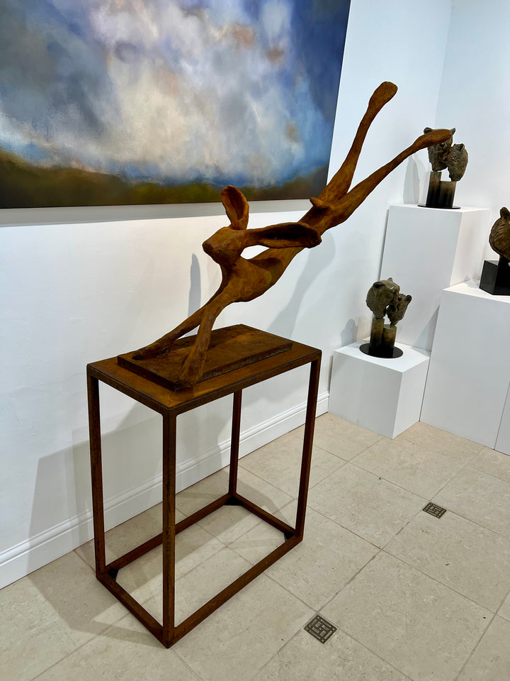 Freedom Majestic Hare sculpture by Min Reid, dynamic leaping hare captured mid-flight in warm bronze toned iron resin, mounted on a wooden base with countryside landscape, green fields and blue sky with white clouds in the background