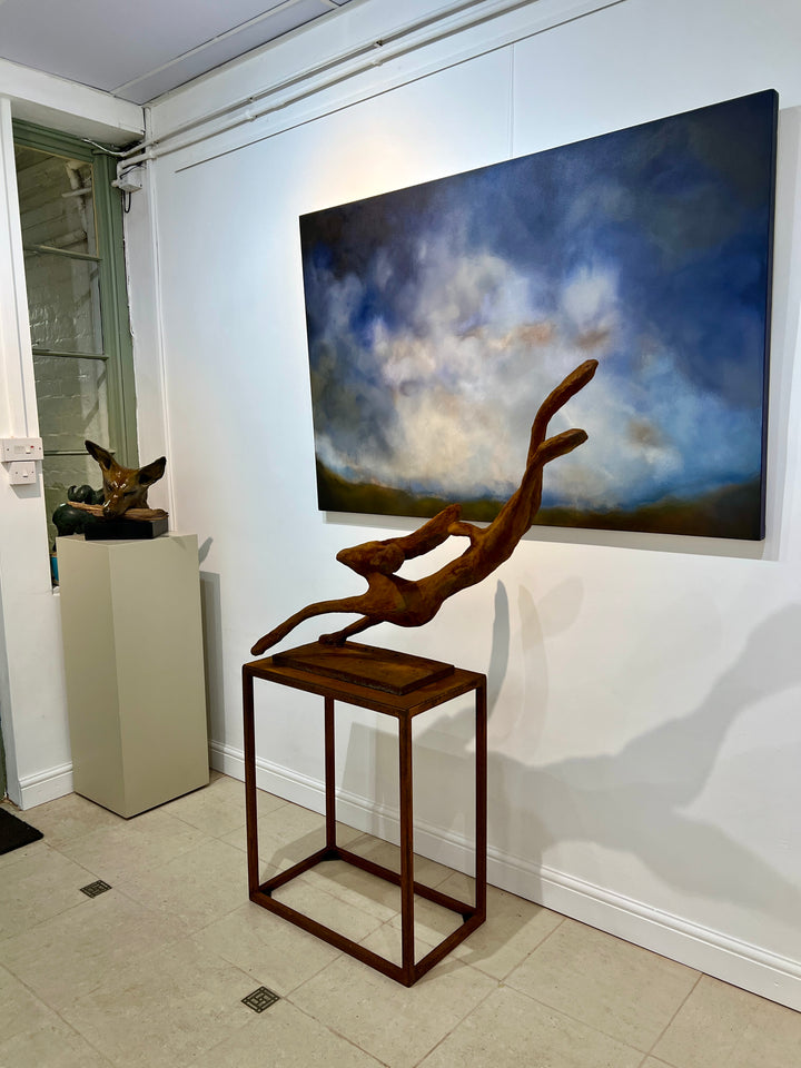 Freedom Majestic Hare sculpture by Min Reid, dynamic leaping hare captured mid-flight in warm bronze toned iron resin, mounted on a wooden base with countryside landscape, green fields and blue sky with white clouds in the background
