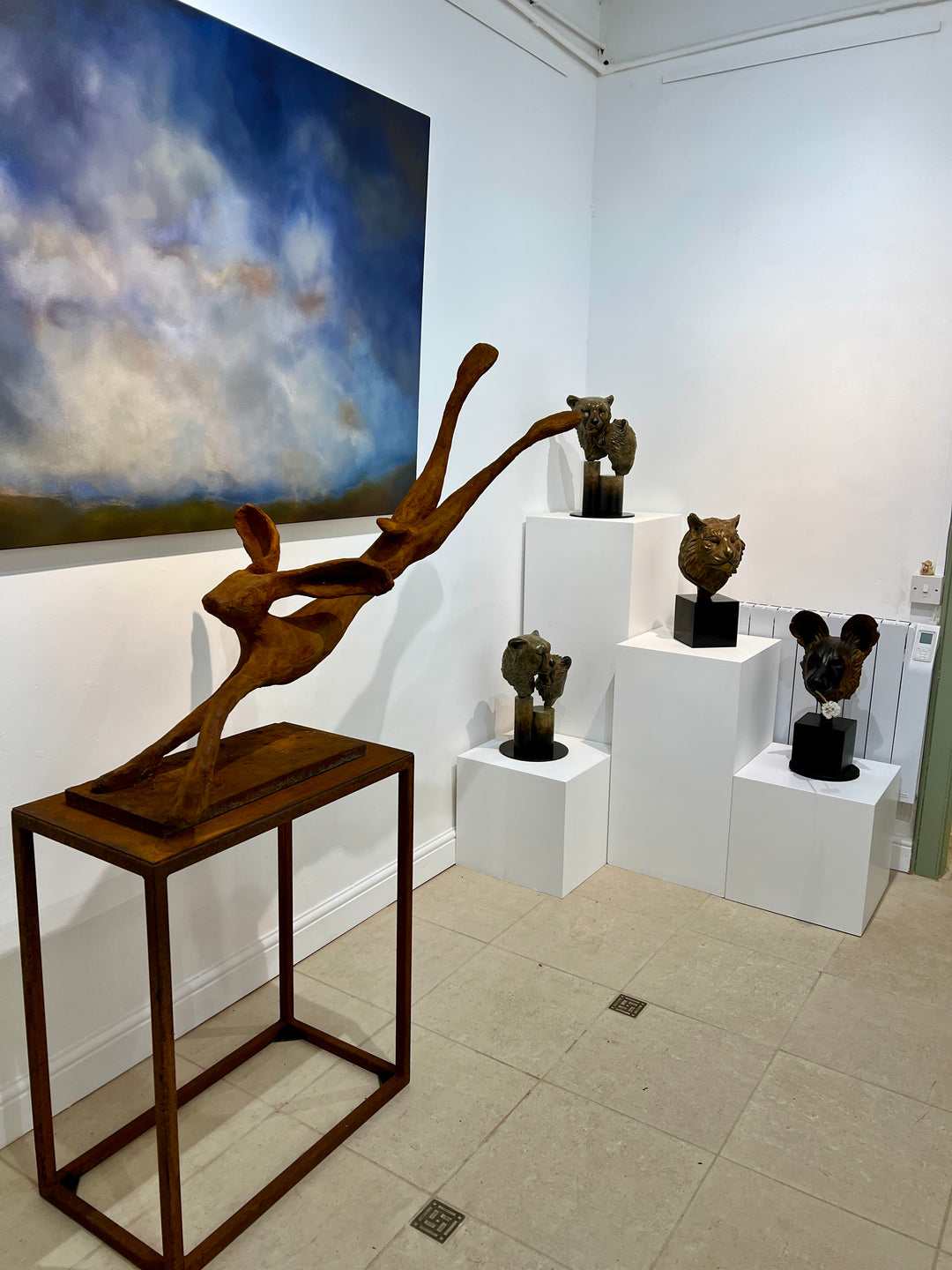 Freedom Majestic Hare sculpture by Min Reid, dynamic leaping hare captured mid-flight in warm bronze toned iron resin, mounted on a wooden base with countryside landscape, green fields and blue sky with white clouds in the background