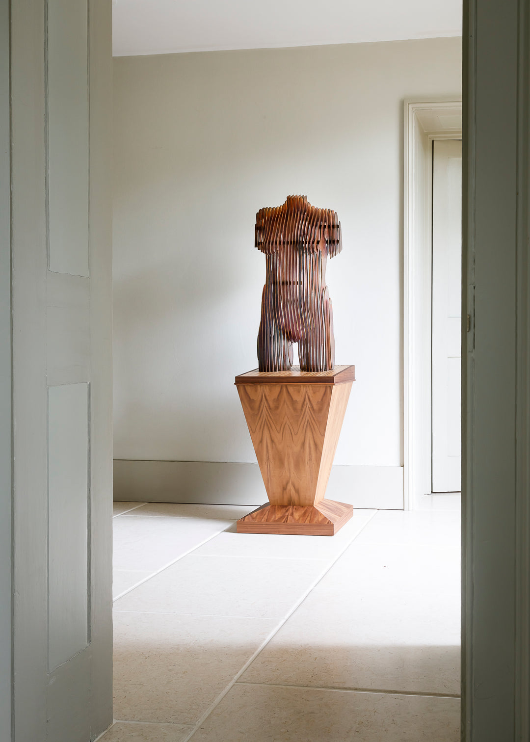 Collaborative sculpture by Tom Hiscox and Fred Baier featuring a stylised female torso formed from layered corten steel and copper plate, mounted on a geometric American walnut and oak base, shown in a light-filled interior setting.