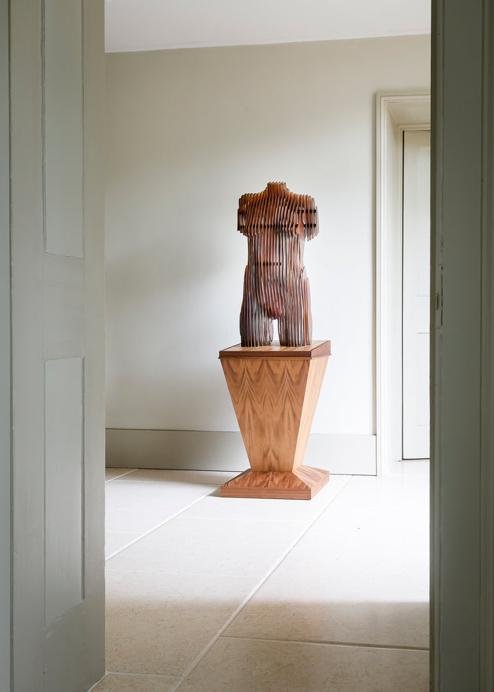 Collaborative sculpture by Tom Hiscox and Fred Baier featuring a stylised female torso formed from layered corten steel and copper plate, mounted on a geometric American walnut and oak base, shown in a light-filled interior setting.