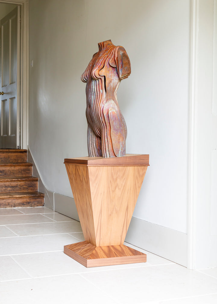 Collaborative sculpture by Tom Hiscox and Fred Baier featuring a stylised female torso formed from layered corten steel and copper plate, mounted on a geometric American walnut and oak base, shown in a light-filled interior setting.