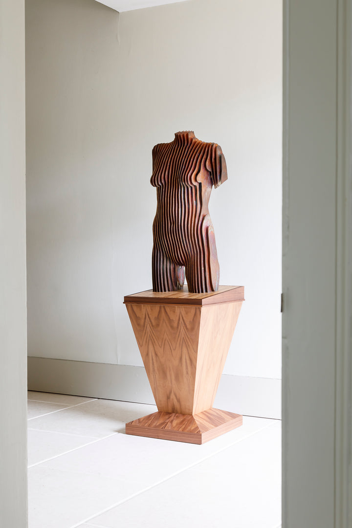 Collaborative sculpture by Tom Hiscox and Fred Baier featuring a stylised female torso formed from layered corten steel and copper plate, mounted on a geometric American walnut and oak base, shown in a light-filled interior setting.