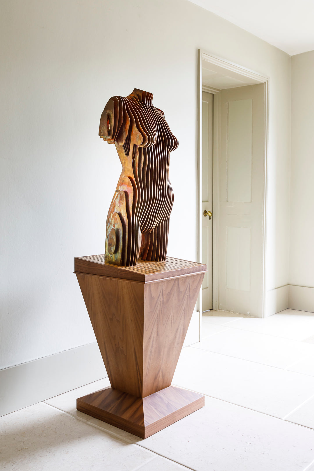 Collaborative sculpture by Tom Hiscox and Fred Baier featuring a stylised female torso formed from layered corten steel and copper plate, mounted on a geometric American walnut and oak base, shown in a light-filled interior setting.