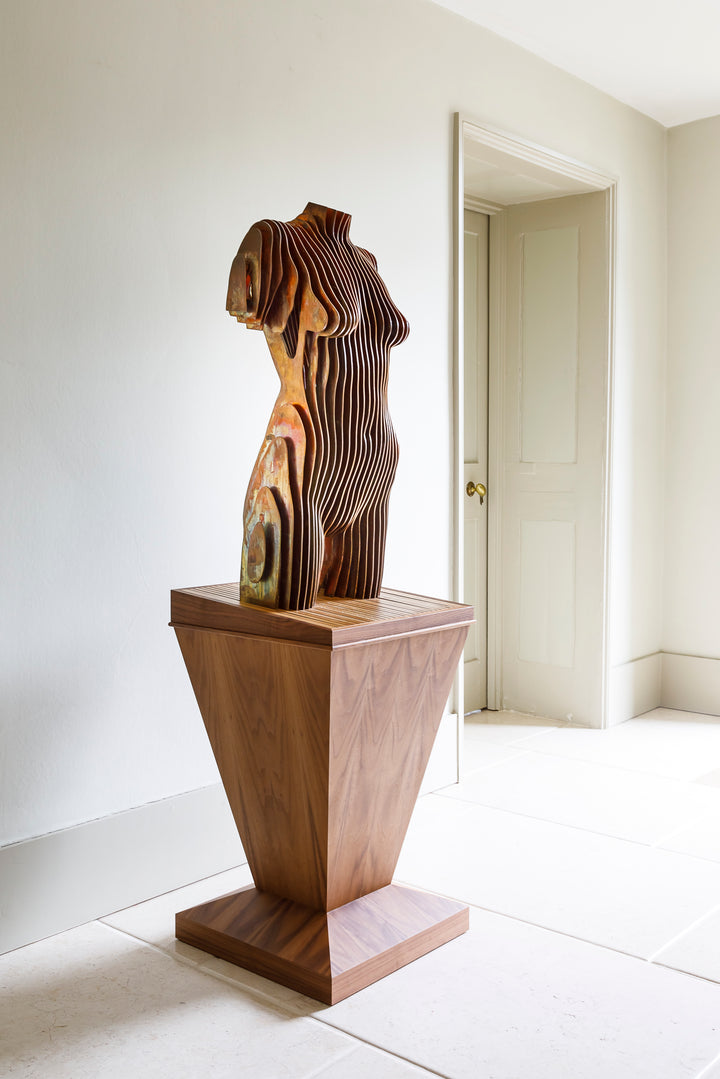 Collaborative sculpture by Tom Hiscox and Fred Baier featuring a stylised female torso formed from layered corten steel and copper plate, mounted on a geometric American walnut and oak base, shown in a light-filled interior setting.