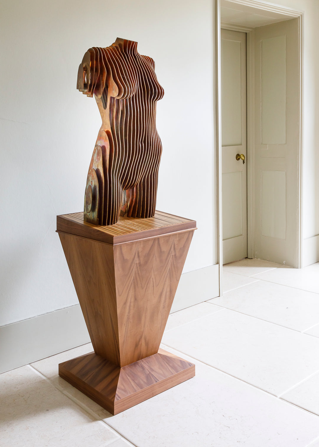 Collaborative sculpture by Tom Hiscox and Fred Baier featuring a stylised female torso formed from layered corten steel and copper plate, mounted on a geometric American walnut and oak base, shown in a light-filled interior setting.