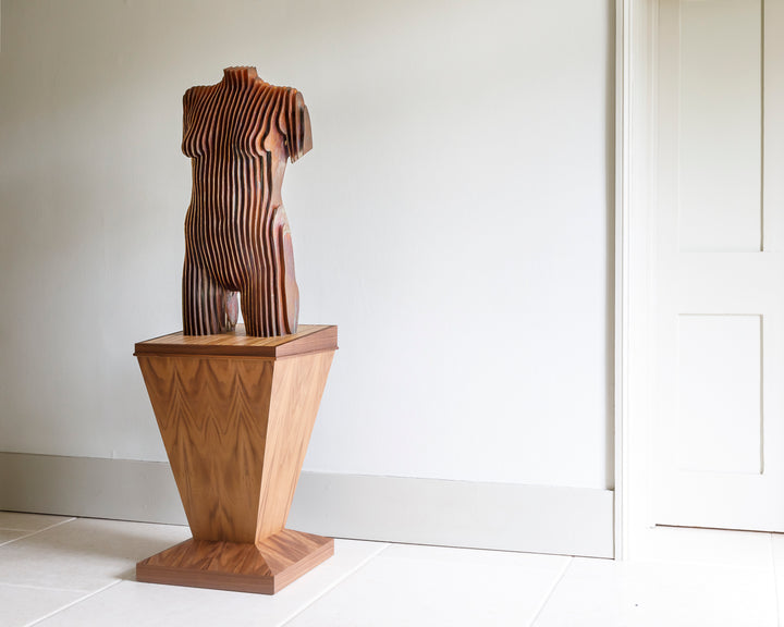 Collaborative sculpture by Tom Hiscox and Fred Baier featuring a stylised female torso formed from layered corten steel and copper plate, mounted on a geometric American walnut and oak base, shown in a light-filled interior setting.