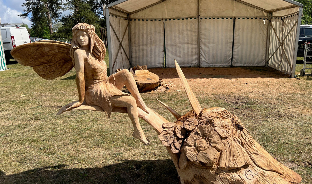 Hand-carved wooden sculpture of a fairy with long flowing hair, flower crown, and textured wings, seated gracefully on a carved branch at an outdoor event.