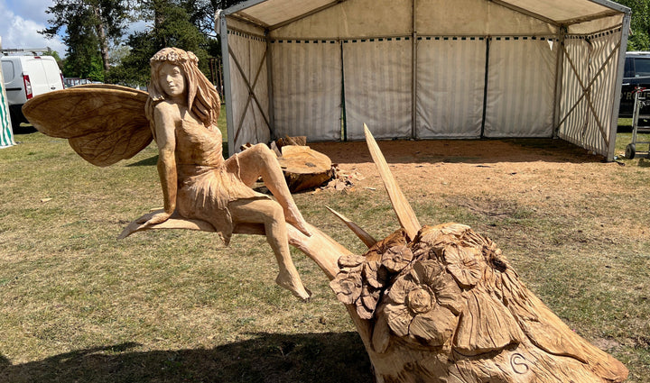 Hand-carved wooden sculpture of a fairy with long flowing hair, flower crown, and textured wings, seated gracefully on a carved branch at an outdoor event.
