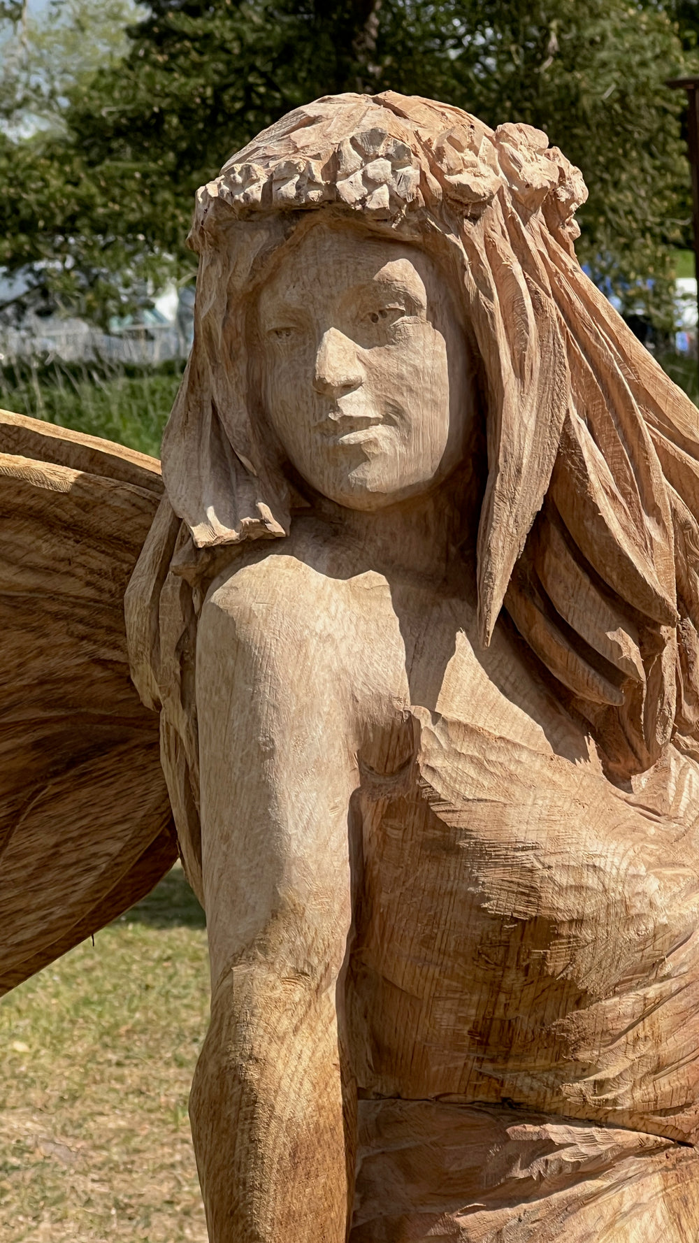 Hand-carved wooden sculpture of a fairy with long flowing hair, flower crown, and textured wings, seated gracefully on a carved branch at an outdoor event.