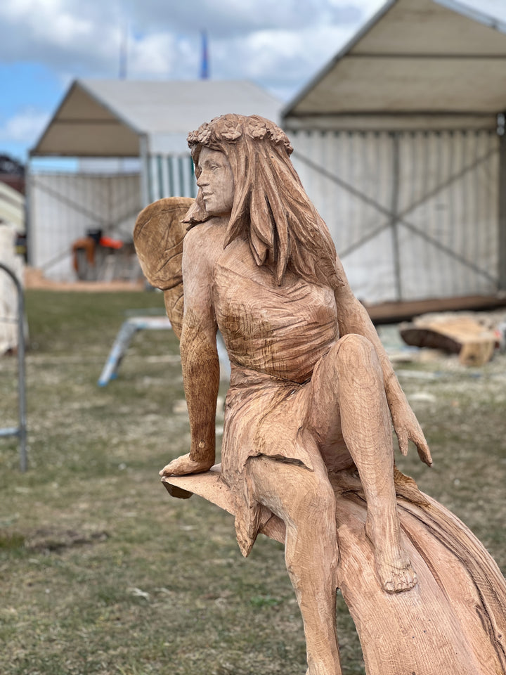 Hand-carved wooden sculpture of a fairy with long flowing hair, flower crown, and textured wings, seated gracefully on a carved branch at an outdoor event.