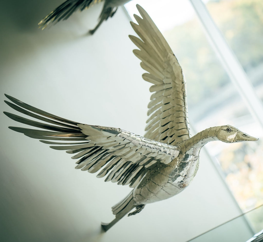 ‘Flying Geese’ by Michael Turner, a unique contemporary stainless steel bird sculpture featuring three geese captured in flight with their wings fully extended. Handcrafted from marine-grade stainless steel, each bird features intricately shaped metallic feathers, detailed wings and a highly polished silver mirror finish. Suspended at different heights within a modern architectural interior, the sculptures create a striking sense of movement, freedom and natural elegance while reflecting the surrounding lig