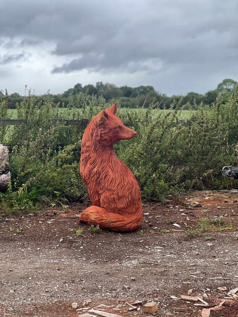 Hand-carved wooden sculpture of a seated fox with its head turned to the right, placed outdoors on rustic ground against a backdrop of overgrown greenery and a moody sky.