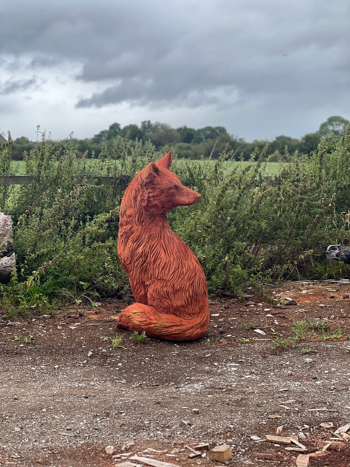 Hand-carved wooden sculpture of a seated fox with its head turned to the right, placed outdoors on rustic ground against a backdrop of overgrown greenery and a moody sky.
