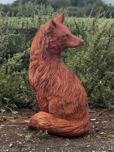 Hand-carved wooden sculpture of a seated fox with its head turned to the right, placed outdoors on rustic ground against a backdrop of overgrown greenery and a moody sky.