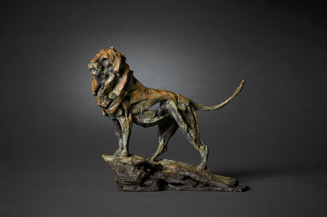 Limited edition bronze lion sculpture by British wildlife sculptor Fred Gordon. The lion stands tall and proud on a rocky base, mane flowing with strong sculptural texture. The bronze surface is richly patinated in earthy tones of deep brown, green, ochre, and subtle hints of red, creating a sense of power and majesty. The pose captures the lion in a moment of still vigilance, tail curved upwards, evoking strength, dignity, and natural authority.