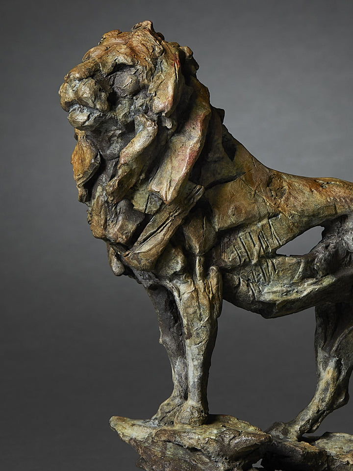 Limited edition bronze lion sculpture by British wildlife sculptor Fred Gordon. The lion stands tall and proud on a rocky base, mane flowing with strong sculptural texture. The bronze surface is richly patinated in earthy tones of deep brown, green, ochre, and subtle hints of red, creating a sense of power and majesty. The pose captures the lion in a moment of still vigilance, tail curved upwards, evoking strength, dignity, and natural authority.