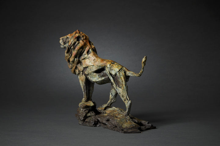 Limited edition bronze lion sculpture by British wildlife sculptor Fred Gordon. The lion stands tall and proud on a rocky base, mane flowing with strong sculptural texture. The bronze surface is richly patinated in earthy tones of deep brown, green, ochre, and subtle hints of red, creating a sense of power and majesty. The pose captures the lion in a moment of still vigilance, tail curved upwards, evoking strength, dignity, and natural authority.