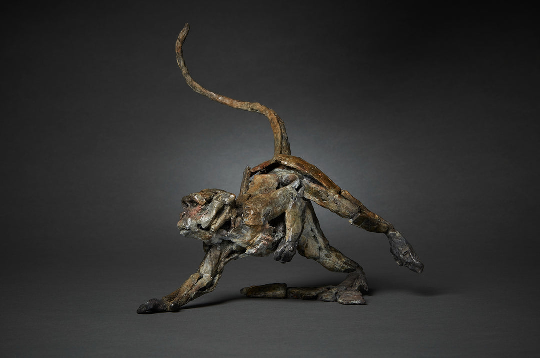 Limited edition bronze sculpture of a running macaque by British sculptor Fred Gordon. The piece captures the primate mid-stride with one arm reaching forward, legs bent in motion, and tail raised in a dynamic arc. The surface is richly textured, with earthy bronze patina tones of brown, black, and green, accented by subtle reddish highlights around the face. A signed and numbered edition of 12, the sculpture embodies movement, vitality, and raw energy in bronze form.