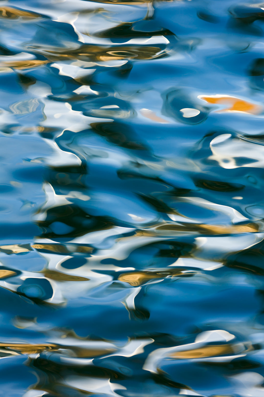 Abstract photograph of light and reflections on gently rippling water, with tones of deep blue, white, and warm golden highlights creating a soft, fluid composition.