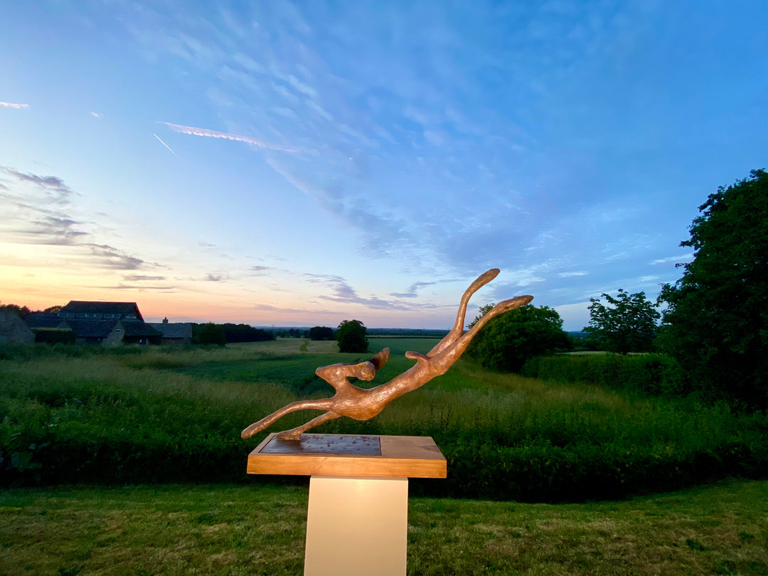 Freedom Majestic Hare sculpture by Min Reid, dynamic leaping hare captured mid-flight in warm bronze toned iron resin, mounted on a wooden base with countryside landscape, green fields and blue sky with white clouds in the background