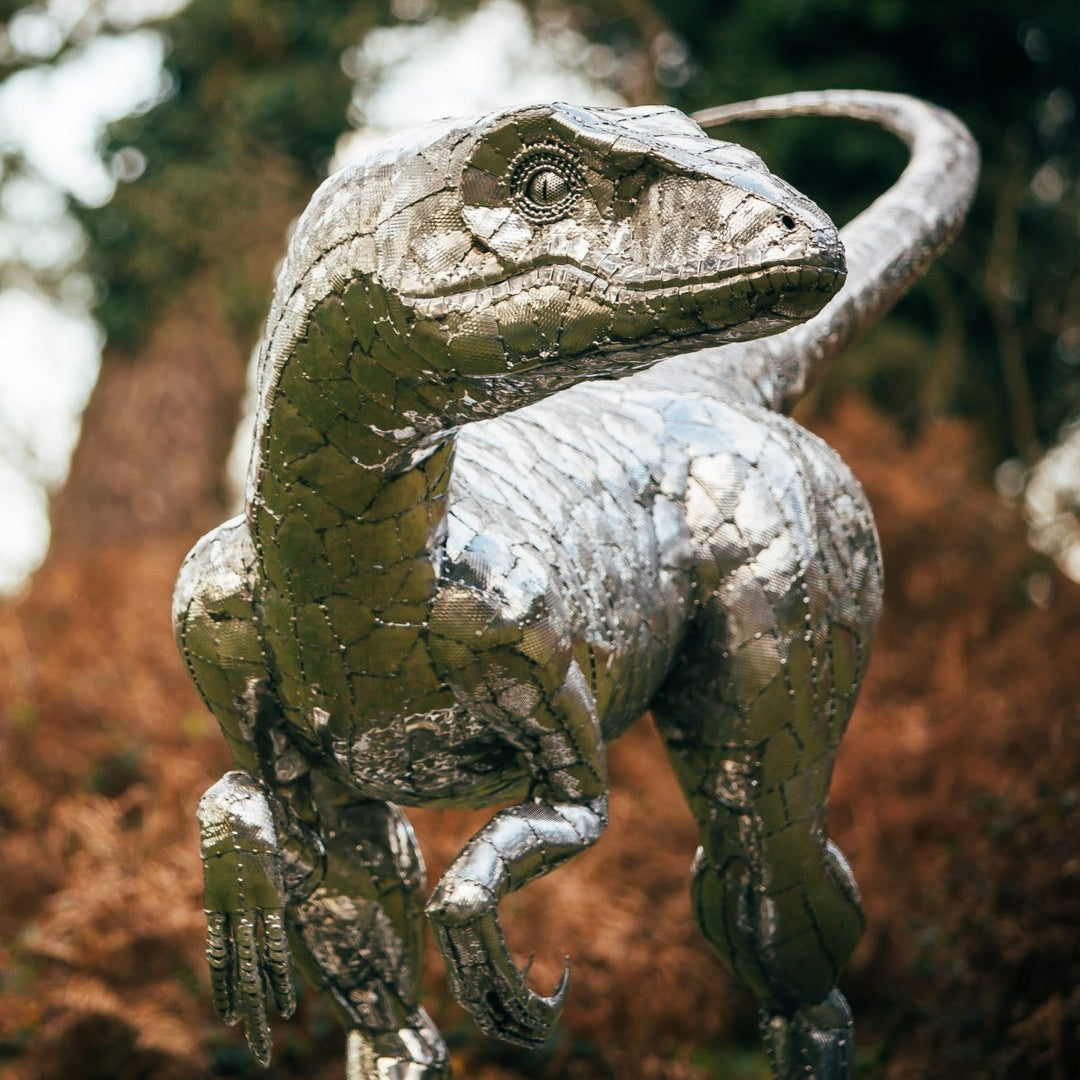 ‘Velociraptor’ by Michael Turner, a unique, one-of-one stainless steel dinosaur sculpture depicting a powerful velociraptor in an alert, dynamic pose. Handcrafted from intricately shaped and welded stainless steel, the sculpture features a polished silver finish, individually detailed metallic scales, muscular hind legs, sharp curved claws, smaller forelimbs and a long, sweeping tail. Standing on a rectangular stainless steel base, this large-scale contemporary sculpture captures the agility and predatory c