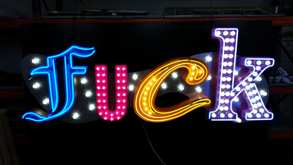 Chris Bracey, neon man, master of glow, gods own junkyard, Illuminated neon sign spelling out an expletive in bold, stylised letters — each letter lit in different colours and styles, including blue script, pink block lights, yellow cursive, and white marquee bulbs, mounted against a dark backdrop.