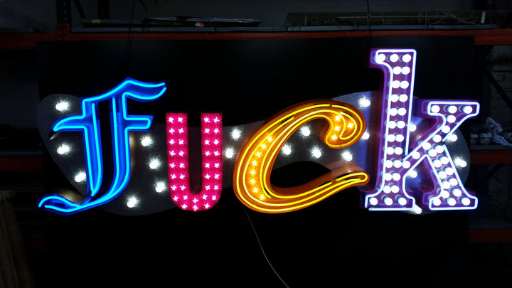 Chris Bracey, neon man, master of glow, gods own junkyard, Illuminated neon sign spelling out an expletive in bold, stylised letters — each letter lit in different colours and styles, including blue script, pink block lights, yellow cursive, and white marquee bulbs, mounted against a dark backdrop.