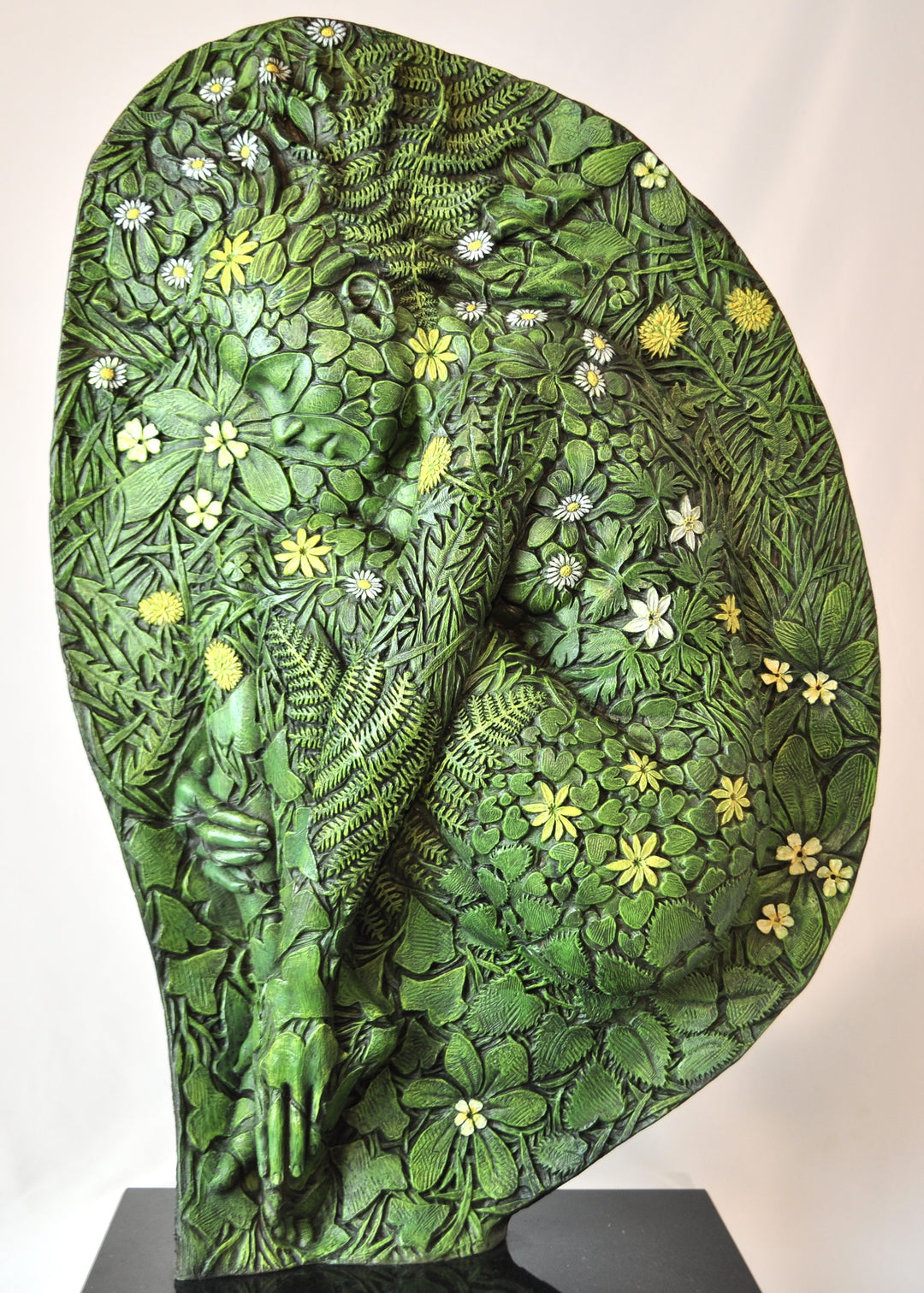 A hand-painted bronze bas-relief sculpture depicting a reclining female form emerging from dense spring foliage, with leaves, ferns and flowers flowing across her body, blending seamlessly into textured earth around the edge.