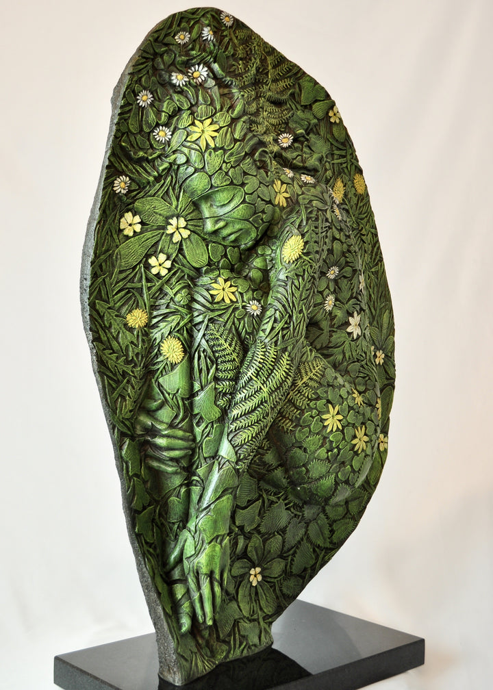 A hand-painted bronze bas-relief sculpture depicting a reclining female form emerging from dense spring foliage, with leaves, ferns and flowers flowing across her body, blending seamlessly into textured earth around the edge.
