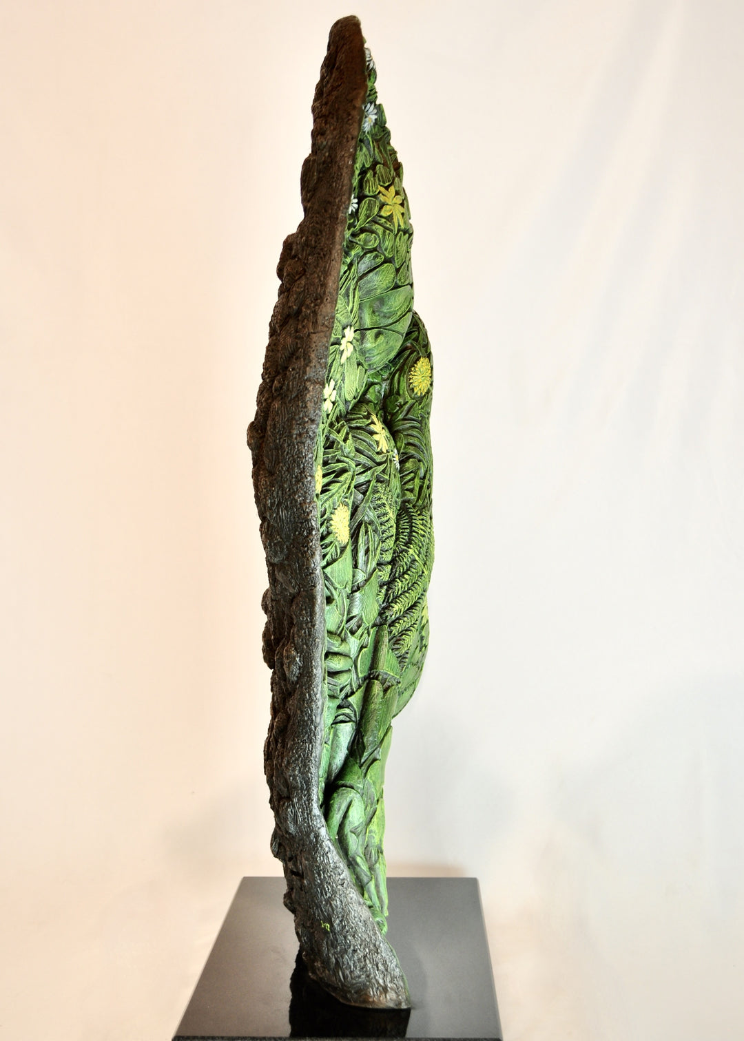 A hand-painted bronze bas-relief sculpture depicting a reclining female form emerging from dense spring foliage, with leaves, ferns and flowers flowing across her body, blending seamlessly into textured earth around the edge.