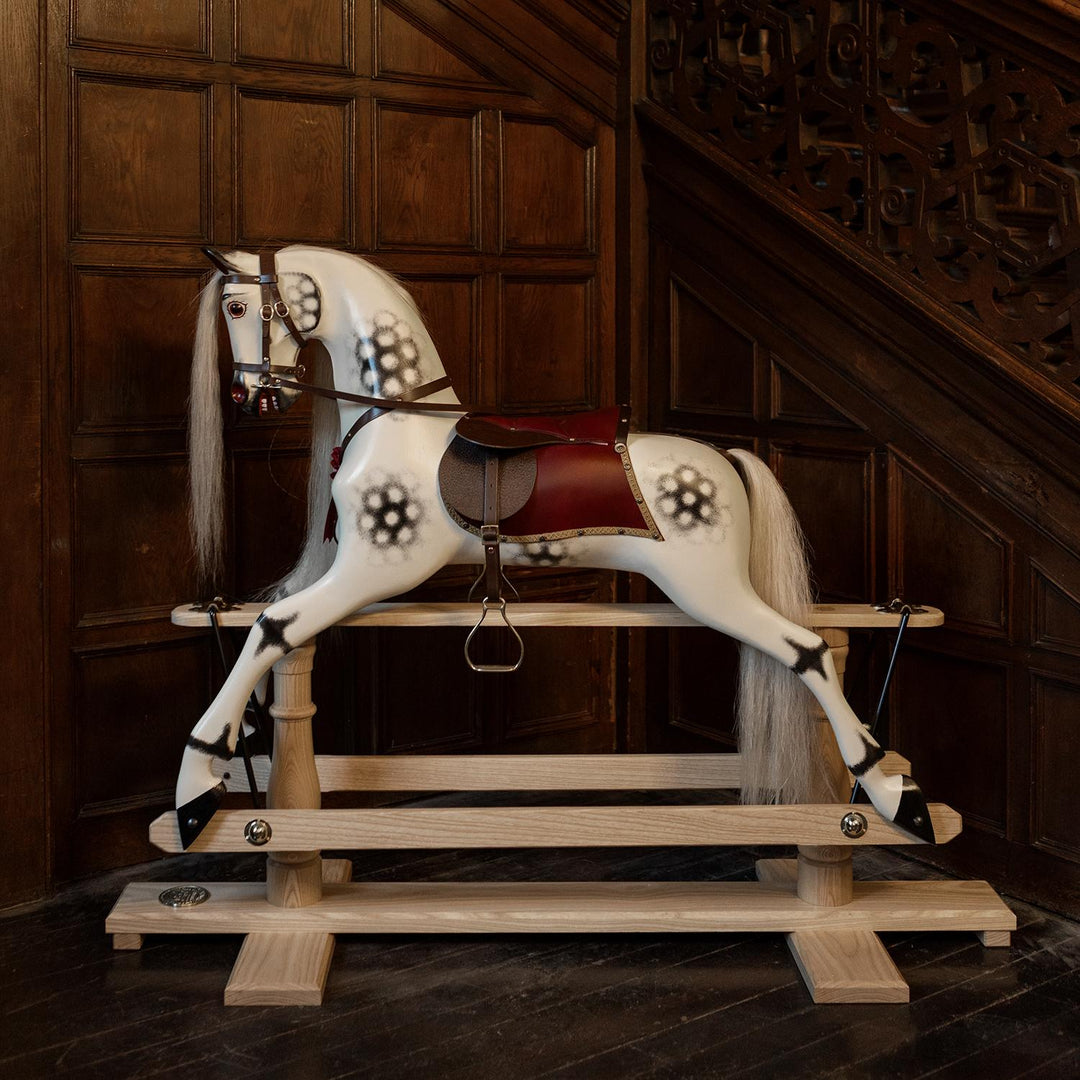 White rocking horse with stylised black dapples, grey mane, and rich burgundy leather tack on a polished oak safety stand.

