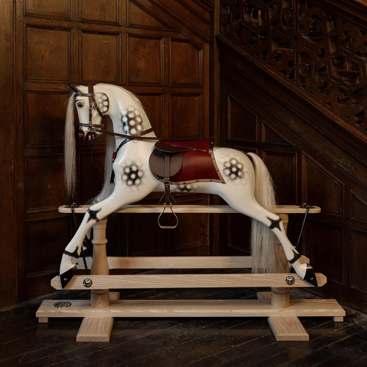 White rocking horse with stylised black dapples, grey mane, and rich burgundy leather tack on a polished oak safety stand.


