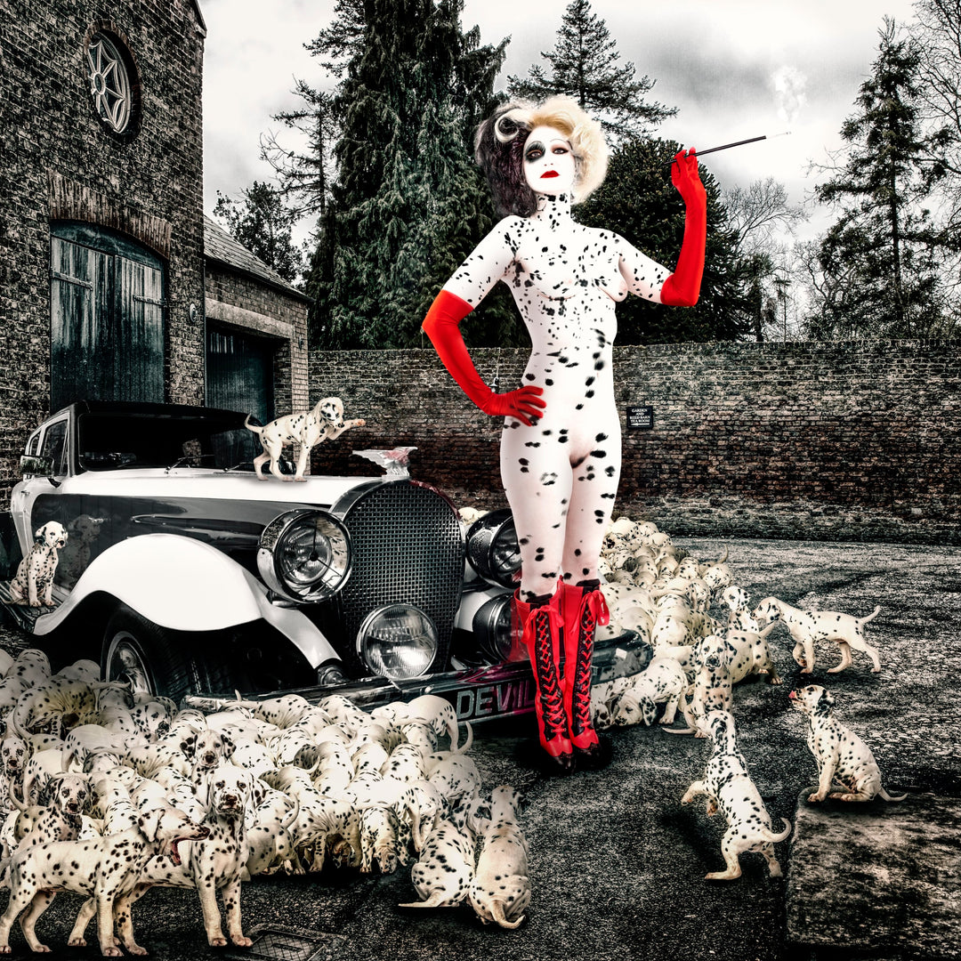 A striking reimagining of Cruella de Vil, portrayed in a bold, nude-toned bodysuit with Dalmatian spots, red gloves, and red lace-up boots, standing confidently in front of a vintage car surrounded by dozens of Dalmatian puppies. Her pose is empowered and commanding, cigarette holder in hand, transforming her from villain to unapologetic leader. The moody backdrop of brick buildings and towering trees frames this cinematic, fashion-forward scene.