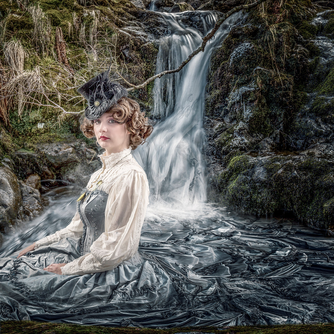 A woman in a Victorian-style gown sits serenely at the base of a waterfall, her silver-blue dress seamlessly transforming into the flowing water beneath her. The scene merges realism with fantasy, blending human presence with nature in an ethereal woodland setting.