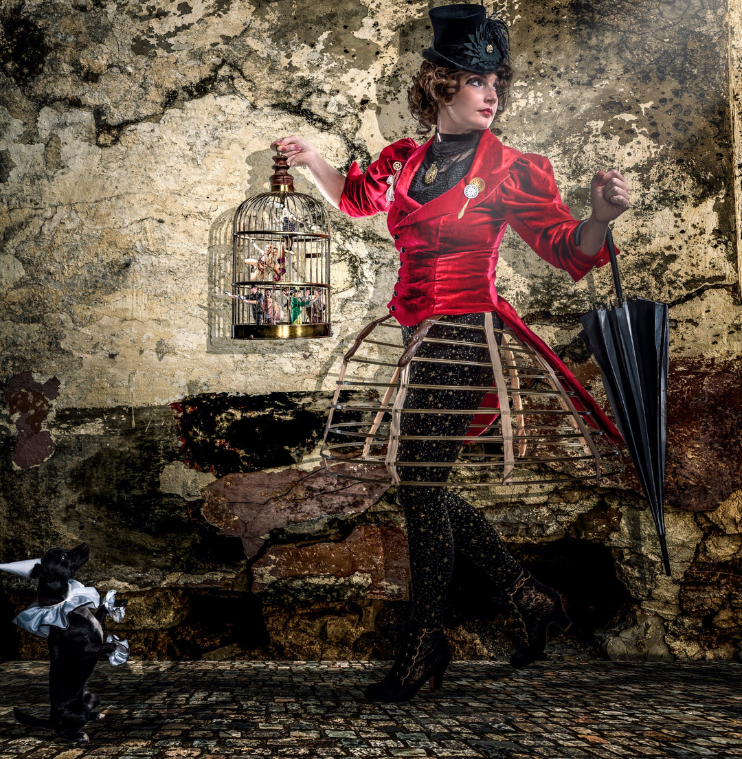 A surreal theatrical composition featuring a woman in a striking red velvet Victorian-style jacket and top hat, holding a black umbrella and a gilded birdcage containing a miniature circus. Her cage-like skirt reveals star-patterned tights, and a small costumed dog gazes up at her. Set against a distressed stone wall, this fantastical image fuses steampunk, performance art, and storytelling.