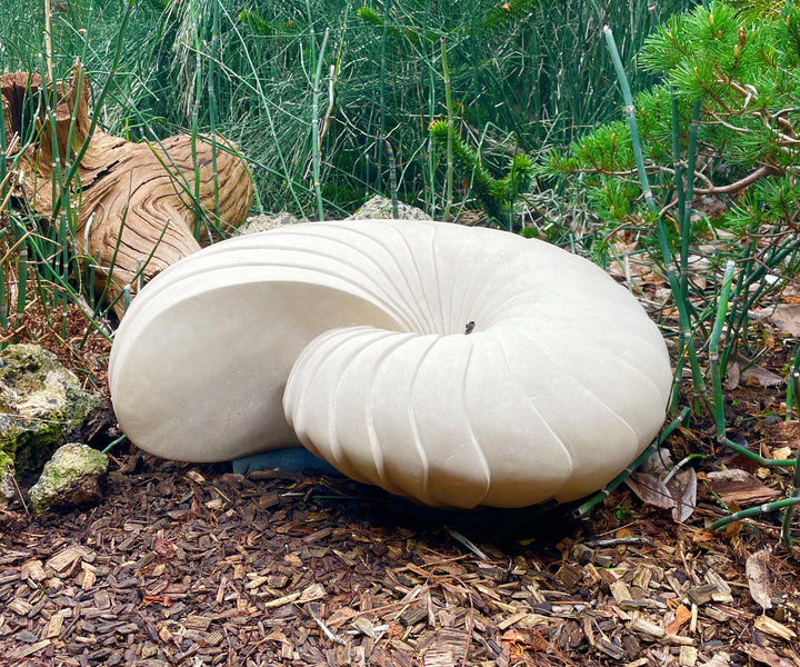 Giant Nautilus by Nicola Axe — large hand-carved Portland stone sculpture shaped like an elegant nautilus shell, smooth pale surface with sweeping ridges, displayed outdoors on a discreet granite base among natural planting.