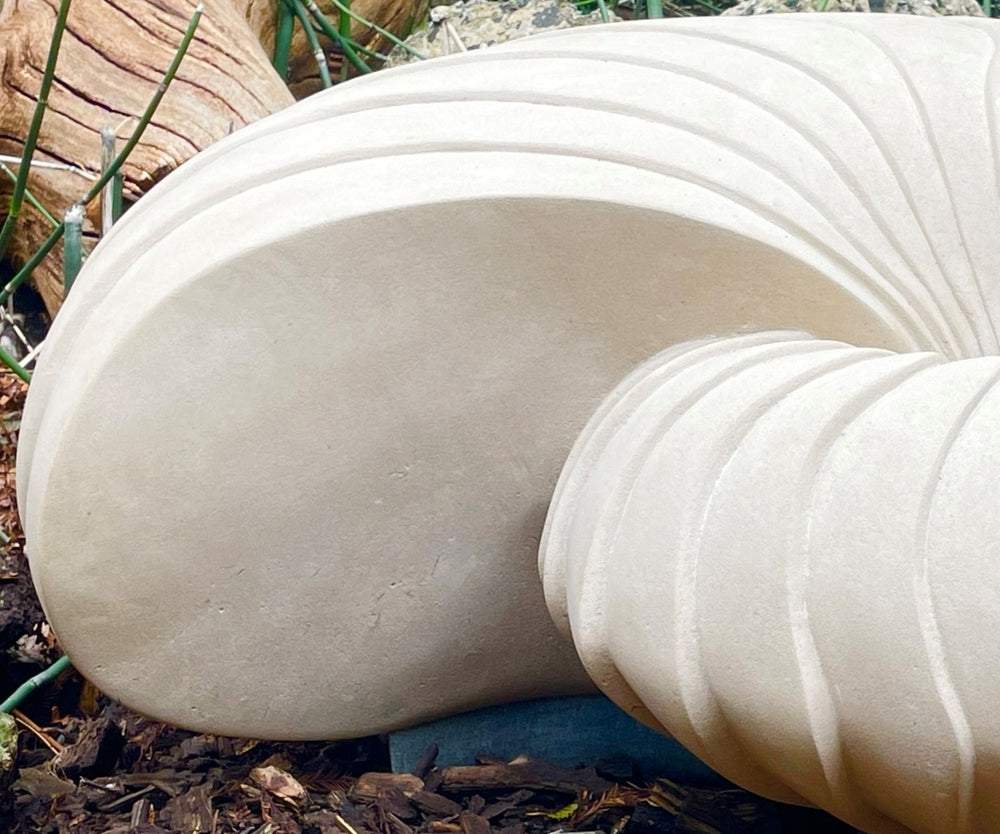 Giant Nautilus by Nicola Axe — large hand-carved Portland stone sculpture shaped like an elegant nautilus shell, smooth pale surface with sweeping ridges, displayed outdoors on a discreet granite base among natural planting.