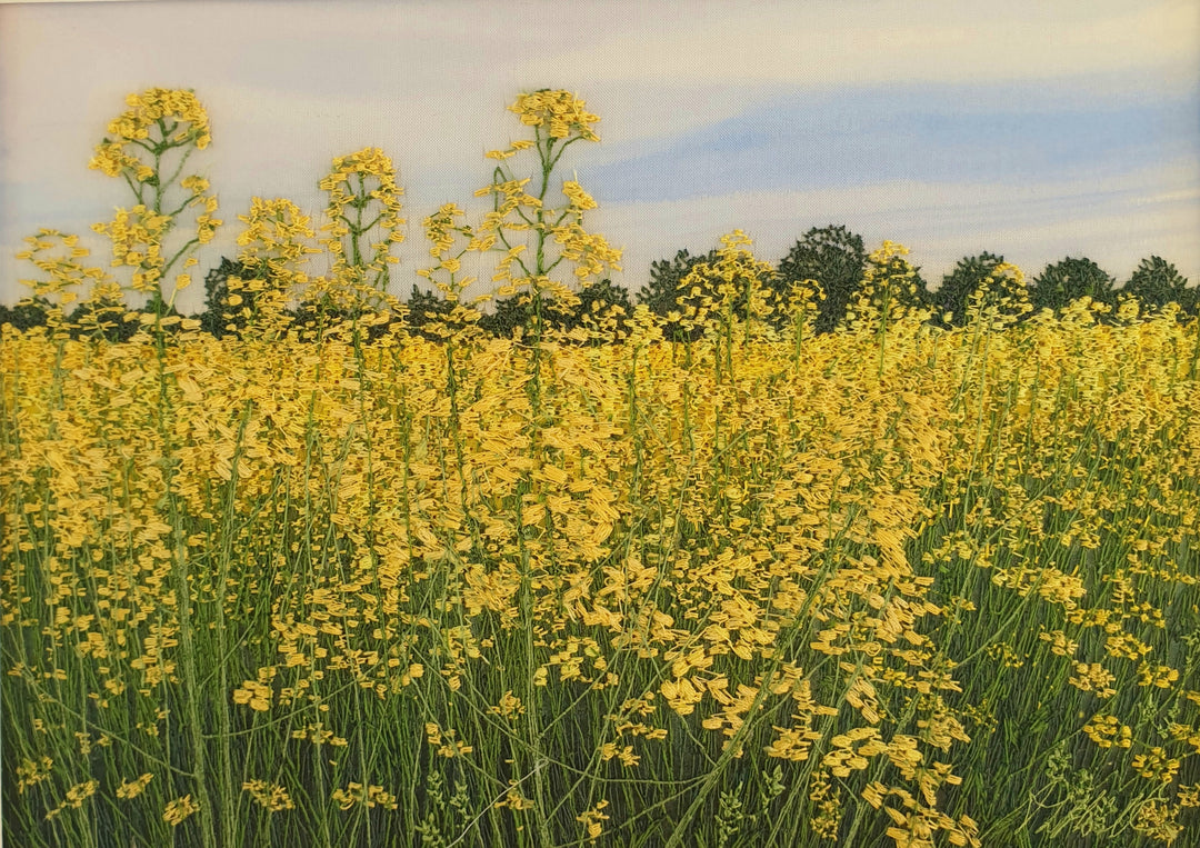 Hand-embroidered field of yellow rapeseed flowers under a soft blue sky, with green trees lining the horizon.

