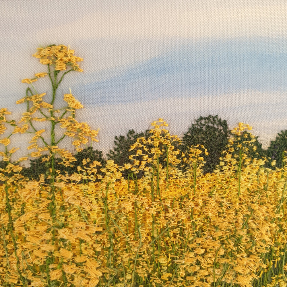 Hand-embroidered field of yellow rapeseed flowers under a soft blue sky, with green trees lining the horizon.

