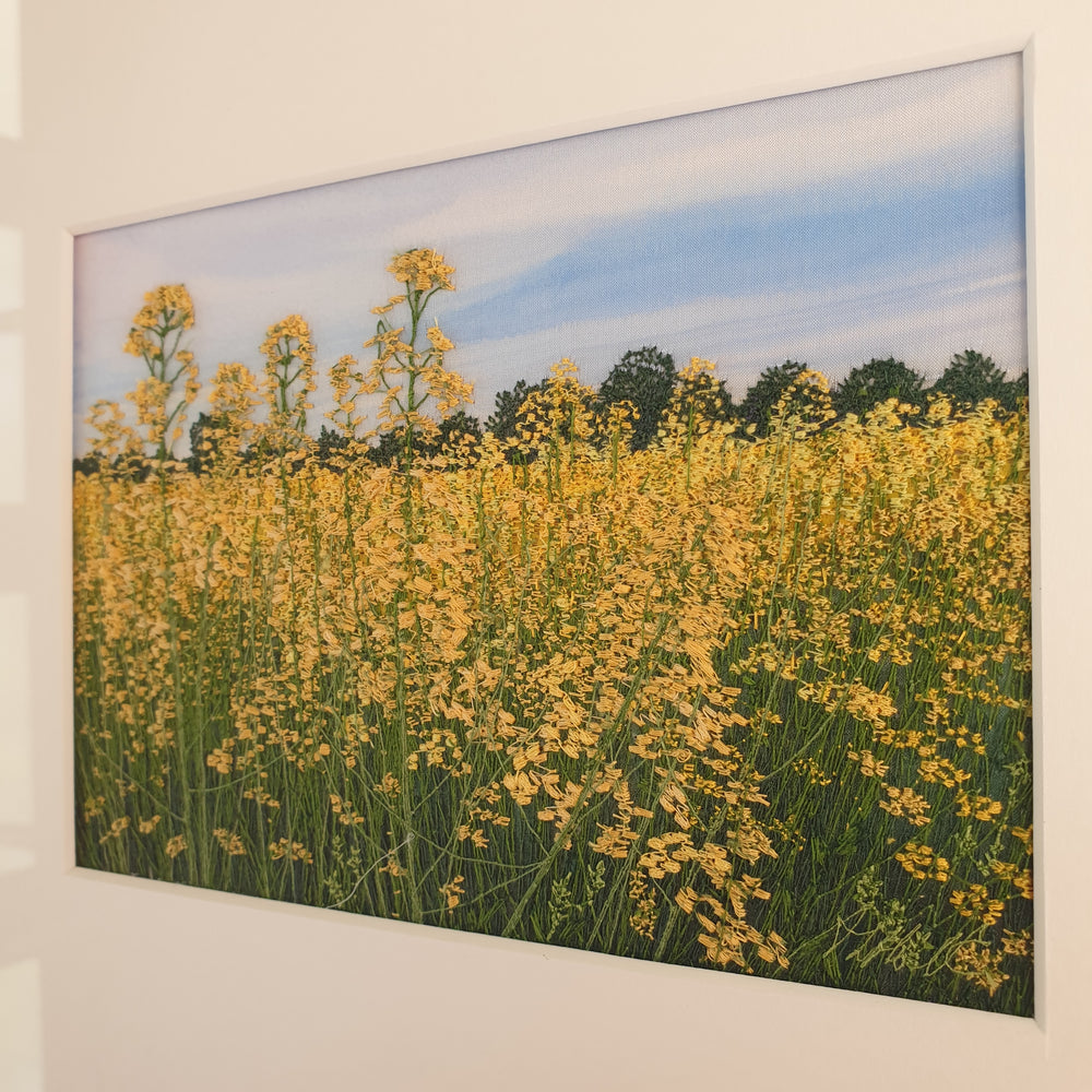 Hand-embroidered field of yellow rapeseed flowers under a soft blue sky, with green trees lining the horizon.

