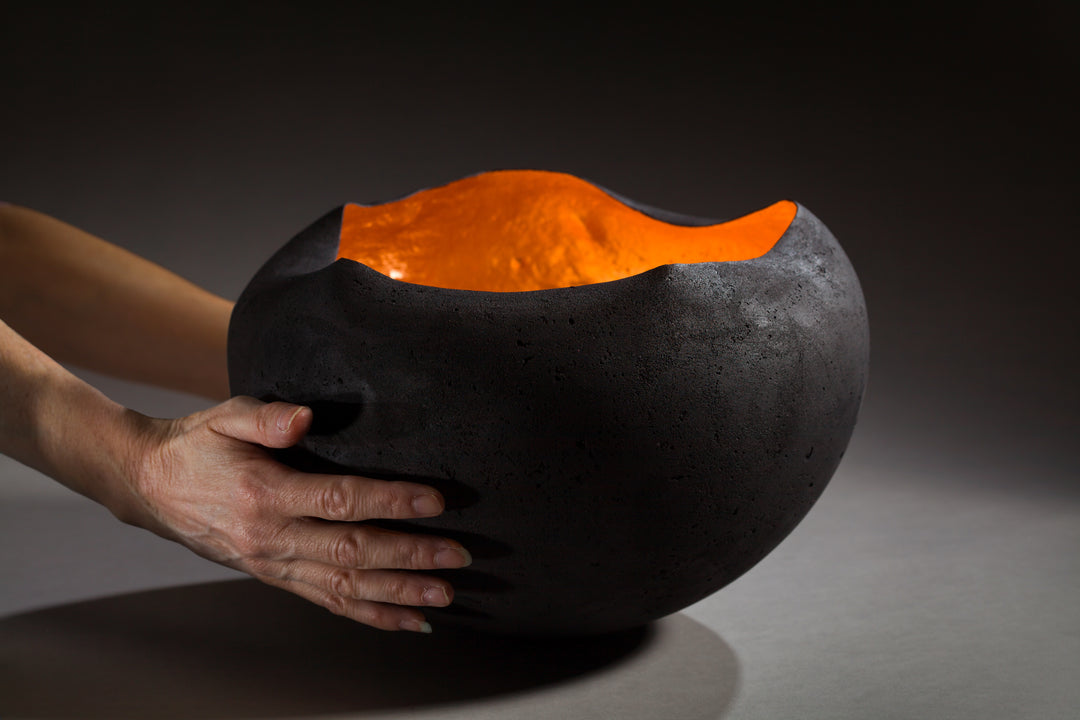 Tracy Nicholls glass sculpture Glow — a hand-formed pâte de verre orb with a rough matte black exterior and glowing copper leaf interior. The vessel radiates warmth, with light reflecting across its curved inner surface, creating the illusion of fire glowing from within.