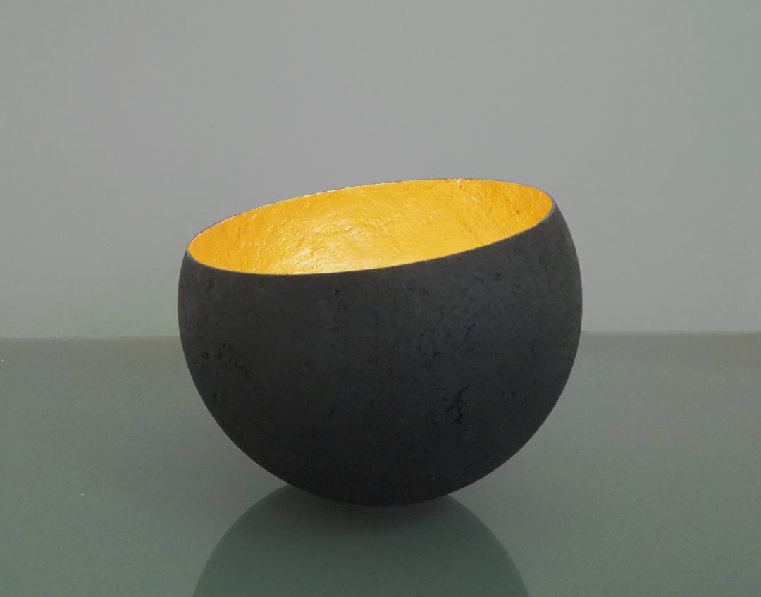 Glow: 22ct Gold by Tracy Nicholls — a one-off kiln-cast glass vessel made using the pâte de verre technique. The piece features a thin, eggshell-like black glass exterior with a rough, volcanic texture, contrasted by a luminous interior lined with 22ct gold leaf. Light reflects across the curved golden surface, creating the illusion of an inner radiance, as though the vessel itself glows from within.
