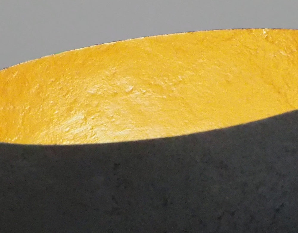 Glow: 22ct Gold by Tracy Nicholls — a one-off kiln-cast glass vessel made using the pâte de verre technique. The piece features a thin, eggshell-like black glass exterior with a rough, volcanic texture, contrasted by a luminous interior lined with 22ct gold leaf. Light reflects across the curved golden surface, creating the illusion of an inner radiance, as though the vessel itself glows from within.