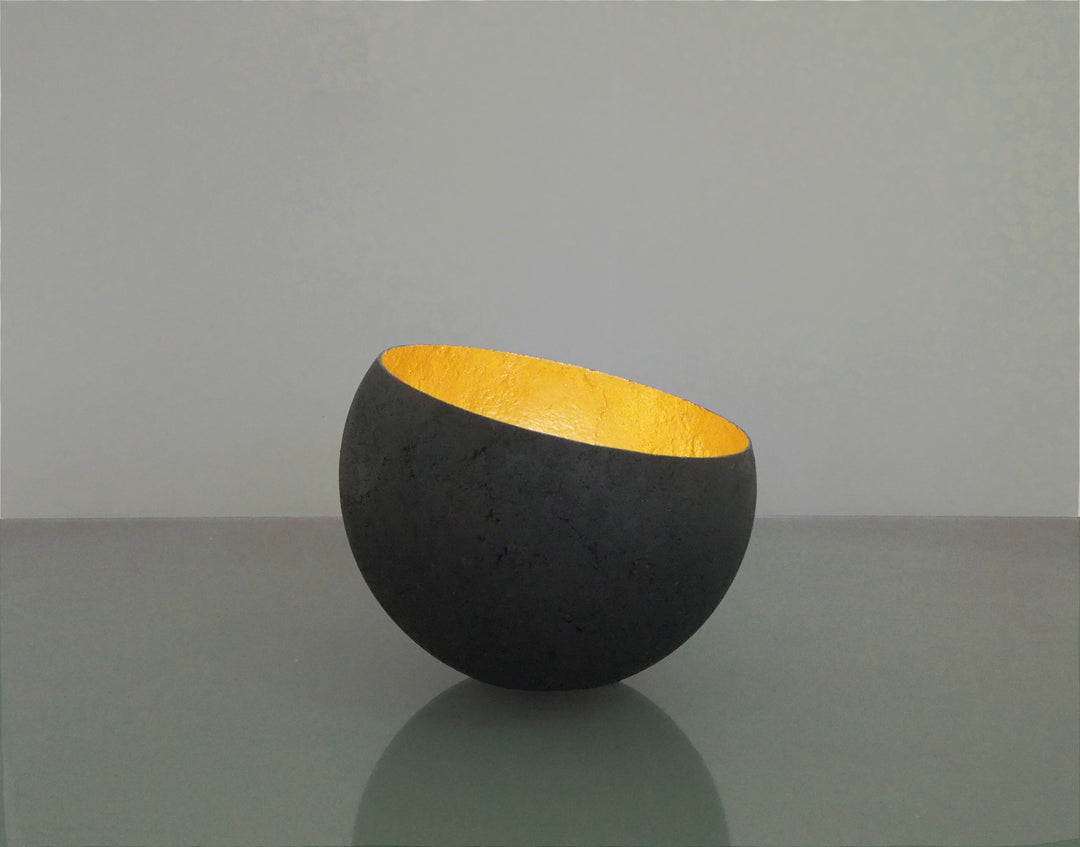 Glow: 22ct Gold by Tracy Nicholls — a one-off kiln-cast glass vessel made using the pâte de verre technique. The piece features a thin, eggshell-like black glass exterior with a rough, volcanic texture, contrasted by a luminous interior lined with 22ct gold leaf. Light reflects across the curved golden surface, creating the illusion of an inner radiance, as though the vessel itself glows from within.
