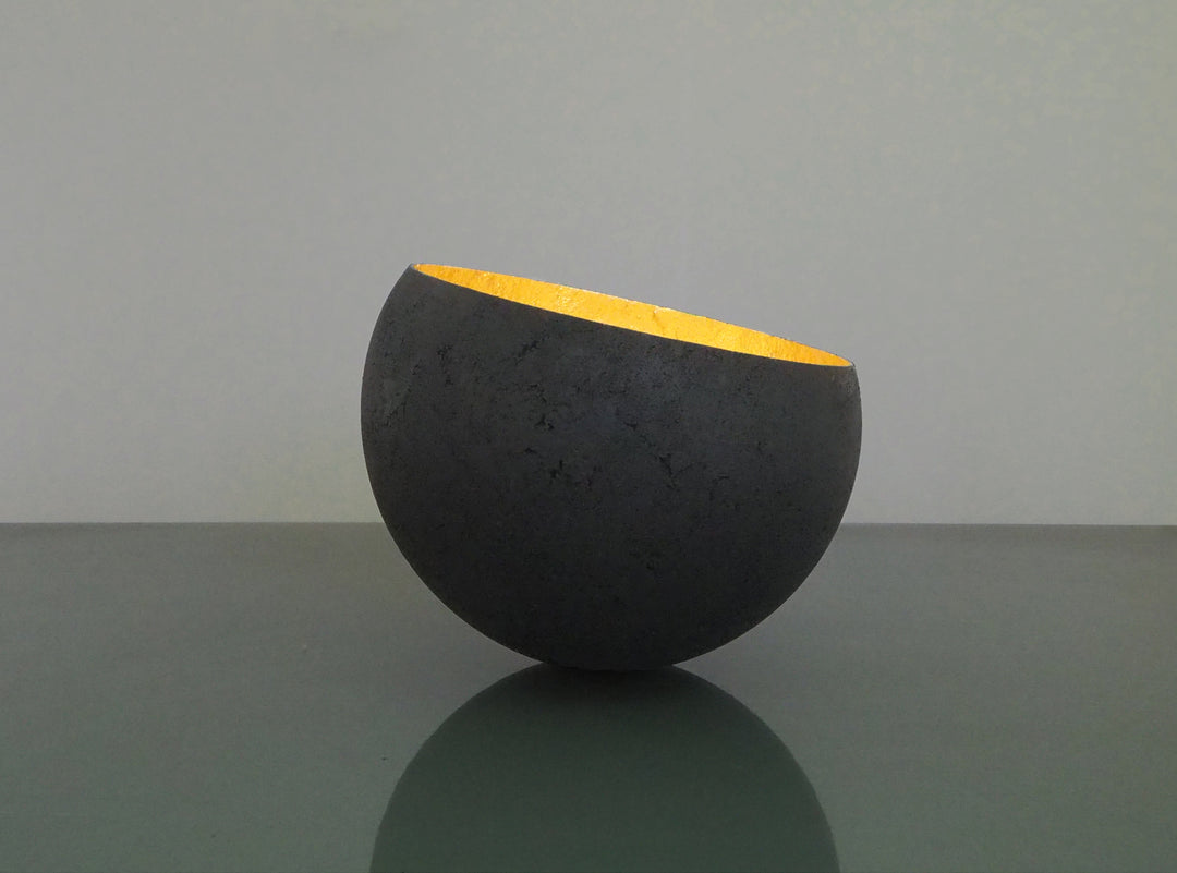 Glow: 22ct Gold by Tracy Nicholls — a one-off kiln-cast glass vessel made using the pâte de verre technique. The piece features a thin, eggshell-like black glass exterior with a rough, volcanic texture, contrasted by a luminous interior lined with 22ct gold leaf. Light reflects across the curved golden surface, creating the illusion of an inner radiance, as though the vessel itself glows from within.