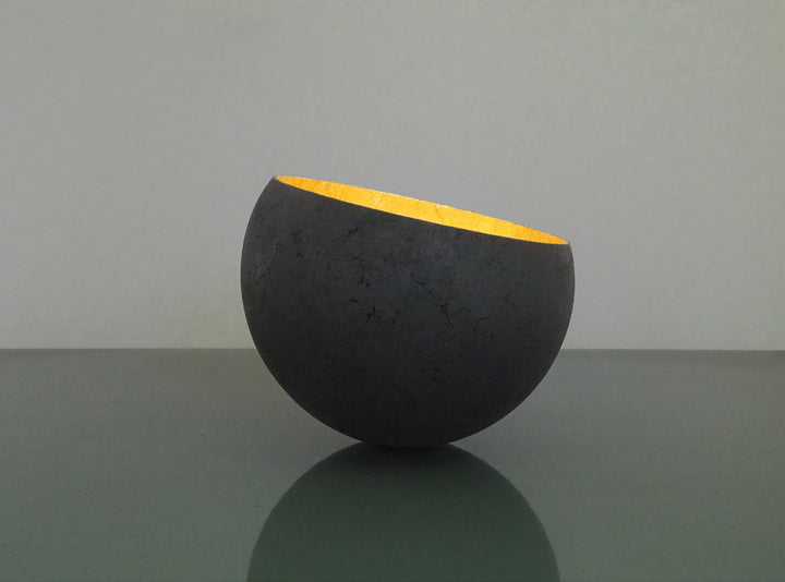 Glow: 22ct Gold by Tracy Nicholls — a one-off kiln-cast glass vessel made using the pâte de verre technique. The piece features a thin, eggshell-like black glass exterior with a rough, volcanic texture, contrasted by a luminous interior lined with 22ct gold leaf. Light reflects across the curved golden surface, creating the illusion of an inner radiance, as though the vessel itself glows from within.