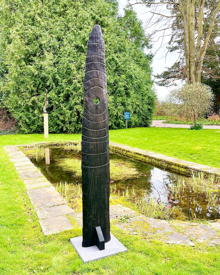 Glen Farrelly sculpture Gobaith, a 205cm tall standing form carved from unwanted oak using the Yakisugi technique, inspired by ancient standing stones. Featuring radiating carved ring-mark patterns and a central circular aperture, the charred blackened surface reveals warm natural oak tones beneath, creating a striking presence of resilience, hope, and renewal in both garden and public spaces.