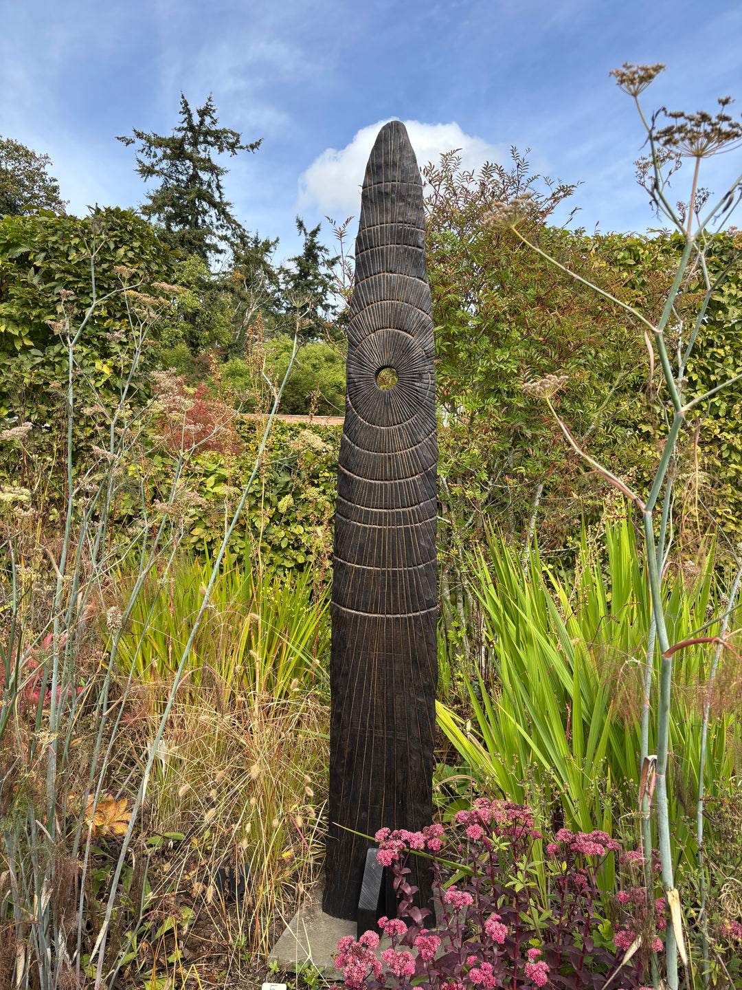 Glen Farrelly sculpture Gobaith, a 205cm tall standing form carved from unwanted oak using the Yakisugi technique, inspired by ancient standing stones. Featuring radiating carved ring-mark patterns and a central circular aperture, the charred blackened surface reveals warm natural oak tones beneath, creating a striking presence of resilience, hope, and renewal in both garden and public spaces.