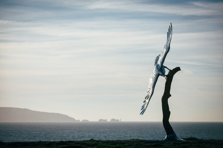 ‘Golden Eagle’ by Michael Turner, a unique, one-of-one stainless steel wildlife sculpture depicting a magnificent golden eagle soaring with its powerful wings fully extended. Handcrafted from polished stainless steel, the sculpture features individually shaped metallic feathers, intricately detailed plumage, a sharply curved beak and striking eyes. Finished in reflective silver with subtle gold tones from the surrounding light, the eagle is mounted on a tall, weathered tree trunk rising from a square metal 